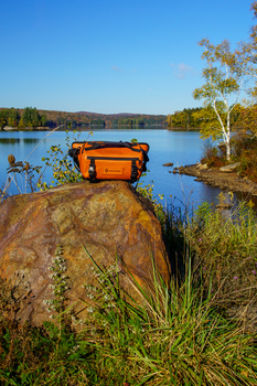Orange waterproof case positioned on a rock overlooking a calm lake with autumn foliage.