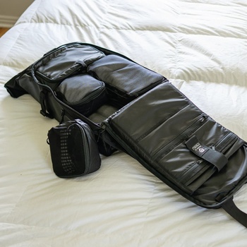 Black camera backpack with padded compartments and attached lens case on white bedding.
