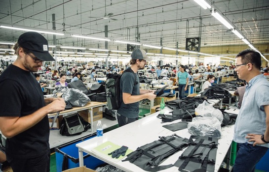 Workers inspecting and sorting garments at tables in a large factory warehouse with industrial ceiling and fluorescent lighting.