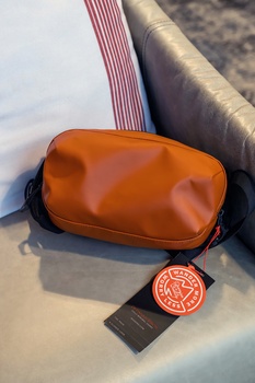 Cognac-colored leather camera bag with black straps displayed on white surface next to circular badge.