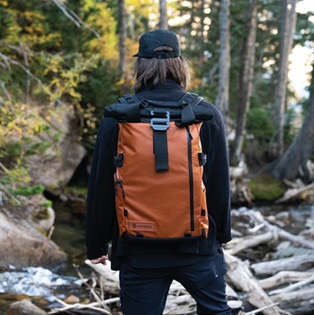 Hiker wearing orange backpack and dark jacket stands on forest trail with fallen logs and evergreen trees.