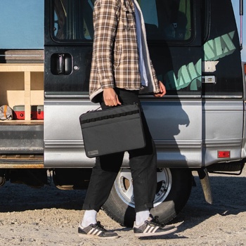 Person exiting a van while holding a black camera case, wearing plaid shirt and black pants.