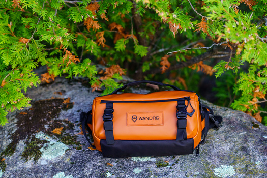 Orange and black waterproof gear bag sitting on a lichen-covered rock surrounded by green moss and foliage.