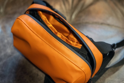 Orange camera bag with black accents and shoulder strap, shown open to display interior lining.