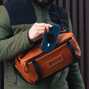 Person holding a smartphone next to an orange camera bag with black straps and zipper details.