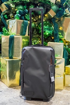 Black rolling luggage positioned in front of gold gift boxes and green holiday foliage backdrop.