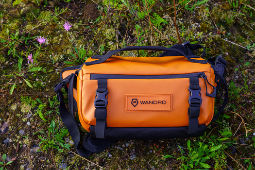 Orange and black waterproof camera bag with shoulder straps sitting on moss and grass.