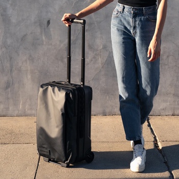 Person in jeans and white sneakers pulling a black rolling suitcase against a textured gray wall.
