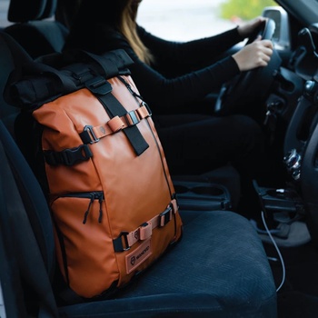 Orange leather backpack secured with straps on car seat next to driver holding steering wheel.