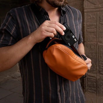 Man holding an orange camera bag while adjusting a mirrorless camera with a striped shirt.