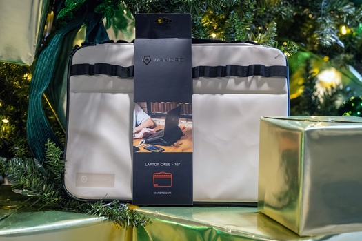 White padded laptop carrying case with black straps and branding displayed on green shelf with decorative foliage.