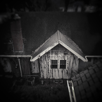 Weathered wooden birdhouse with peaked roof mounted on dark background in black and white.