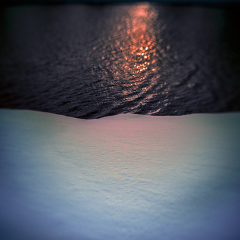 Sunset or sunrise light reflecting off dark water above a snow or ice surface.
