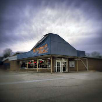 Abandoned commercial building with distinctive peaked roof under dramatic cloudy sky.