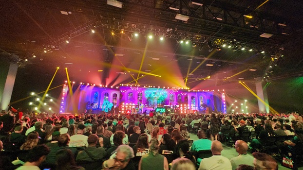 Large concert venue with colorful stage lighting and dense crowd of attendees.
