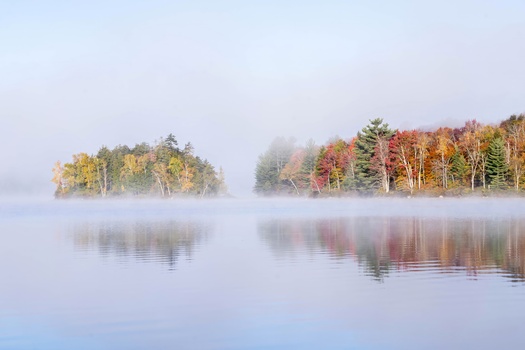 Two forested islands reflected in still lake water surrounded by autumn foliage and morning mist.