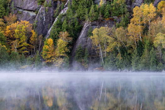 Morning mist rising from a lake with autumn trees and rocky cliffs reflected in still water.
