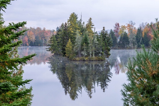 Forested island reflected in still lake water during autumn, with evergreens and colorful deciduous trees.