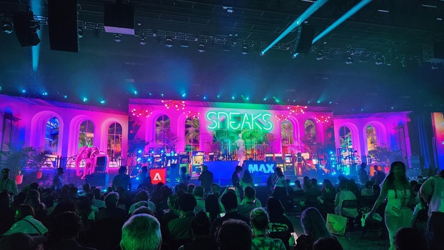 Concert stage with vibrant neon pink and blue lighting, silhouetted crowd, and large green text signage overhead.