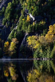 Steep forested mountainside with golden larch trees and a small building nestled among evergreens, reflected in still water below.