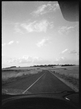 Straight rural road stretching toward horizon under partly cloudy sky, viewed from vehicle dashboard.
