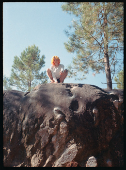 Woman in red sitting atop a large weathered boulder surrounded by pine trees.