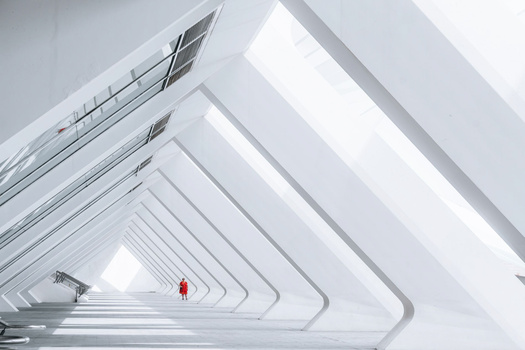 Solitary figure in red standing in a white geometric tunnel with repeating angular frames receding into the distance.