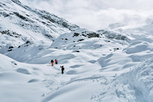 Three hikers traverse a vast snow-covered alpine landscape dotted with small buildings and evergreen trees.
