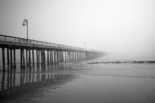 Wooden pier extending into fog with mirror-like wet sand reflections in black and white.