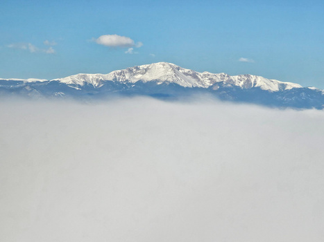 Snow-capped mountain peak rising above a thick blanket of cloud cover under clear blue sky.