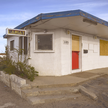 Abandoned roadside structure with curved roof, cream walls, red and yellow doors, and boarded window.