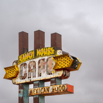 Vintage neon sign for a Mexican food restaurant with yellow lettering and red accents against a cloudy sky.