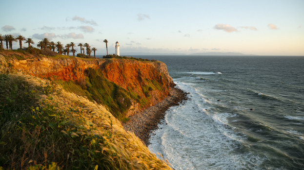 Coastal cliff with lighthouse and palm trees at sunset overlooking the Mediterranean.