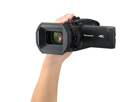 Hand holding a Panasonic 4K camcorder with extended zoom lens and articulating LCD screen.