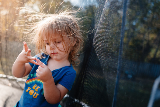 Toddler in blue shirt playing with toy against dark mesh backdrop in bright outdoor sunlight.