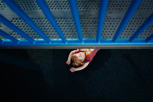 Overhead view of person lying on dark textured floor beneath perforated metal ceiling with blue structural beams.