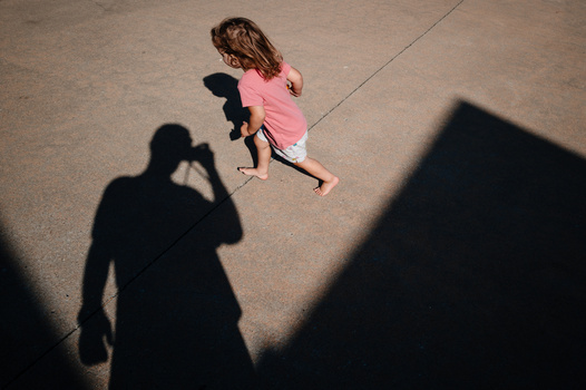 Overhead view of a child in a pink dress running across pavement, with photographer's shadow visible in frame.