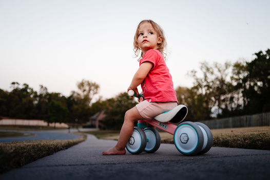 Toddler in pink shirt sitting on blue and white ride-on toy outdoors.
