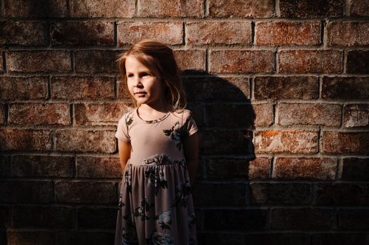 Young girl in a patterned dress standing against a textured brick wall in natural sunlight.
