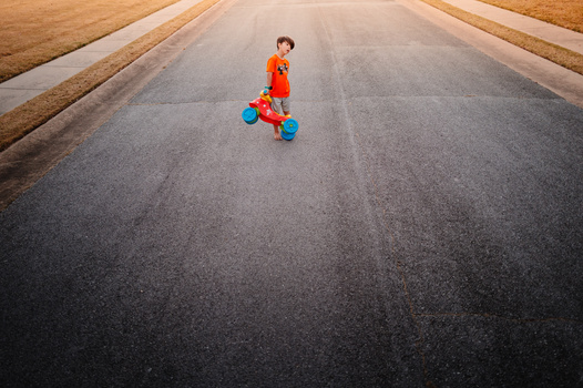 Child in orange clothing riding a toy on a straight asphalt road with fields on either side.