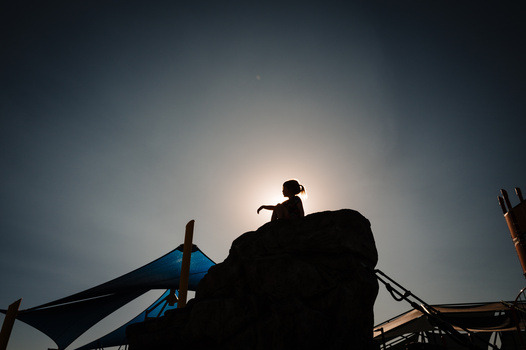Silhouetted child sitting atop a large rock or boulder against a bright sky with climbing equipment visible.
