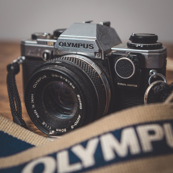 Olympus film SLR camera with 50mm lens on branded camera strap against wooden surface.