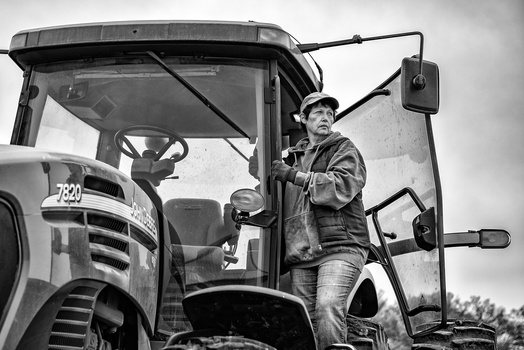 Man standing in cab of vintage truck, wearing hat and work shirt, black and white photograph.