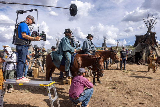 Film crew operating camera equipment on a dirt set with horses and period buildings in background.