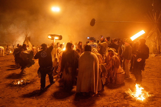 Large nighttime gathering around fires with warm orange lighting and smoke in desert setting.