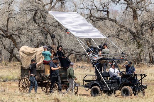 Film production crew setting up a large diffusion panel above vintage wagons in a bare tree grove.