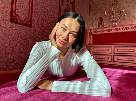 Woman in white ribbed long-sleeve shirt leaning on magenta fabric in ornate red bedroom setting.
