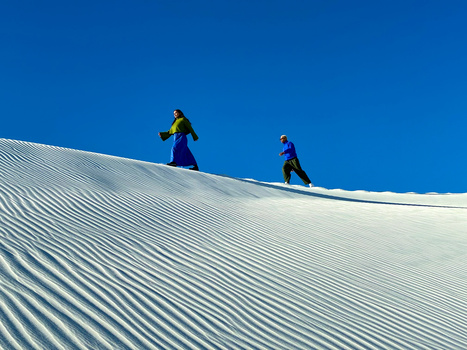 Two hikers ascending a snow-covered dune under clear blue sky, their figures silhouetted against the bright white sand.