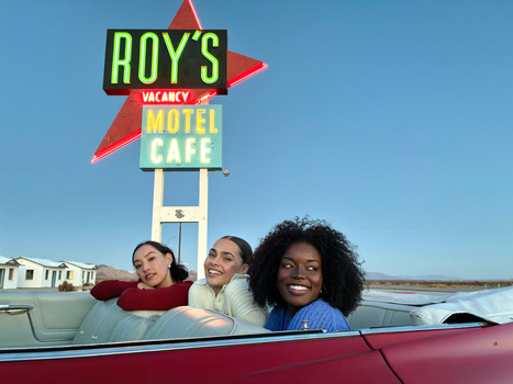 Three young women smiling in a red convertible car with a vintage neon motel sign in the background.