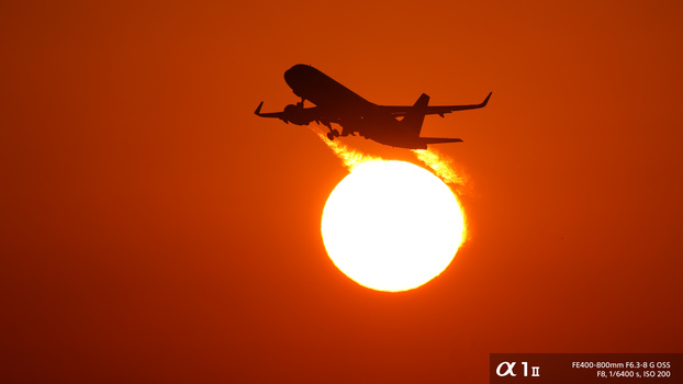 Commercial aircraft silhouetted against a large sun during golden hour, with warm orange sky.